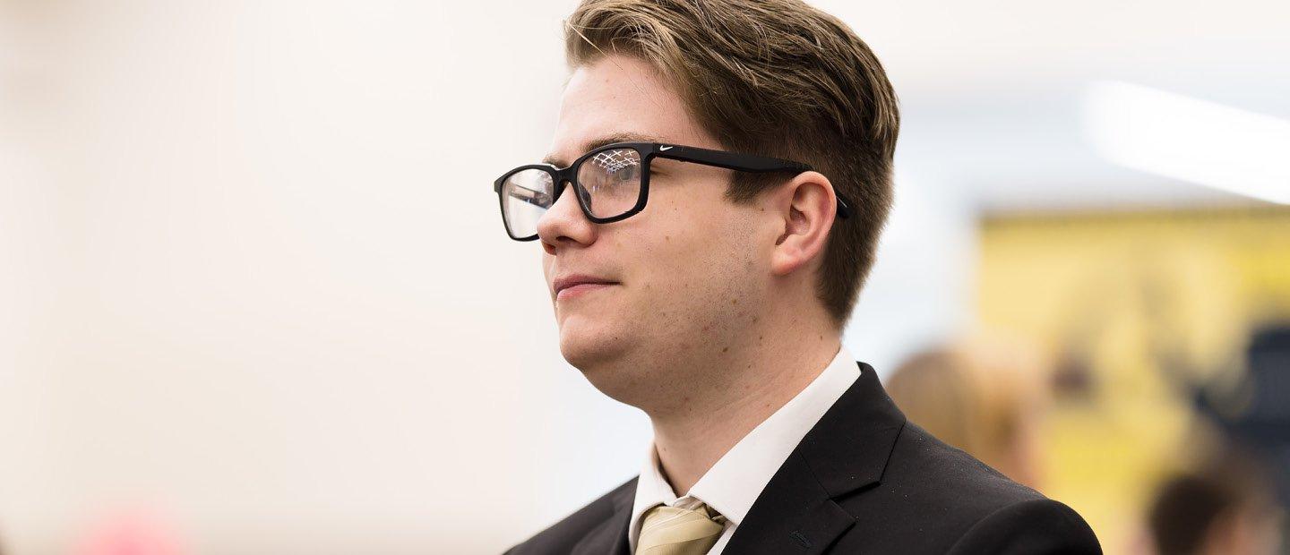 A profile photo of a young man in glasses and a black suit jacket with a gold tie.